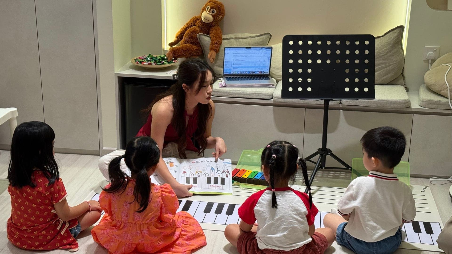 Teacher Jovanna leading our founding Little Stars class, with four preschoolers gathered around a giant floor keyboard