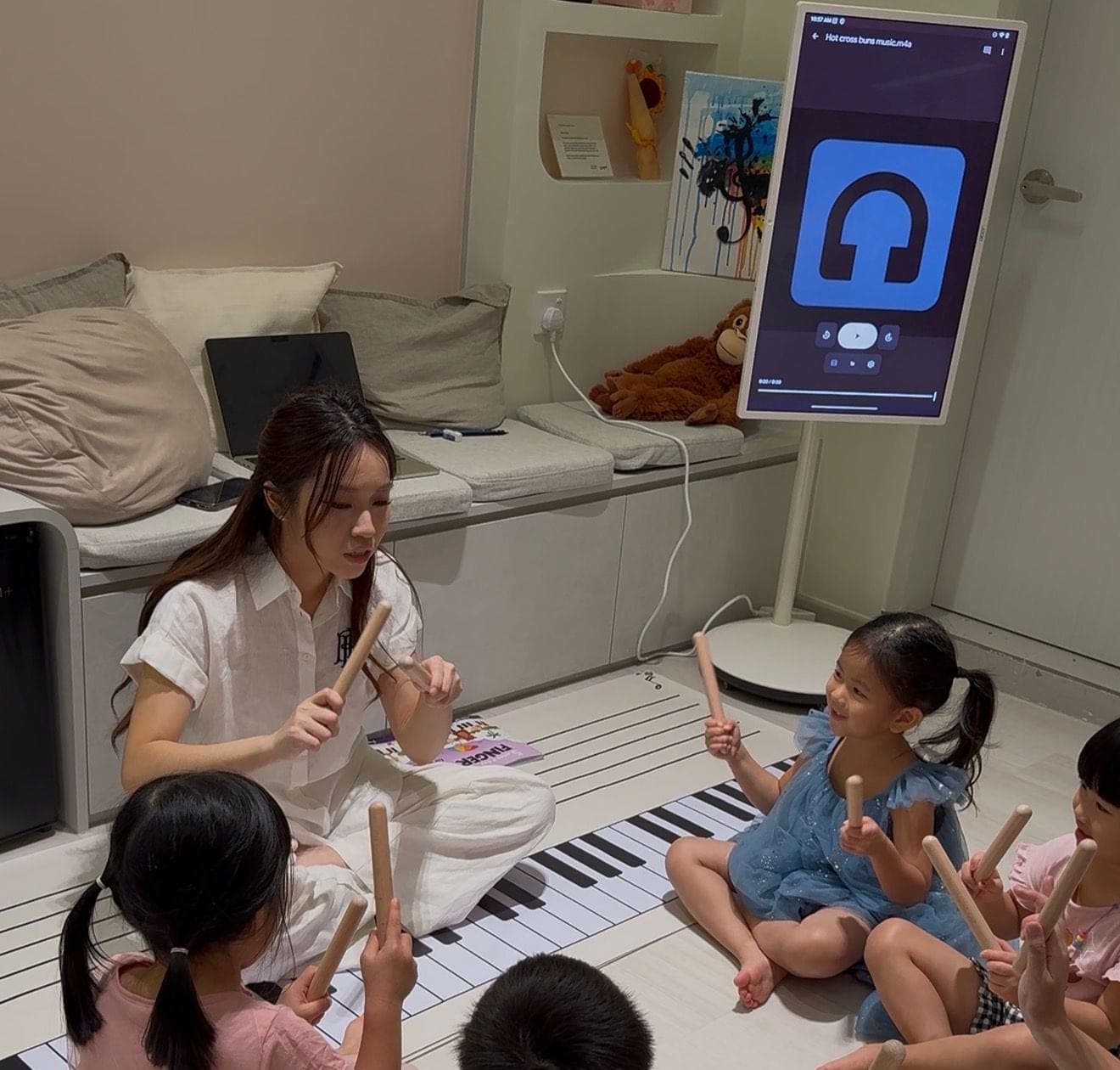 Little Stars students playing rhythm sticks with their teacher around a giant floor keyboard, with Hot Cross Buns on the screen